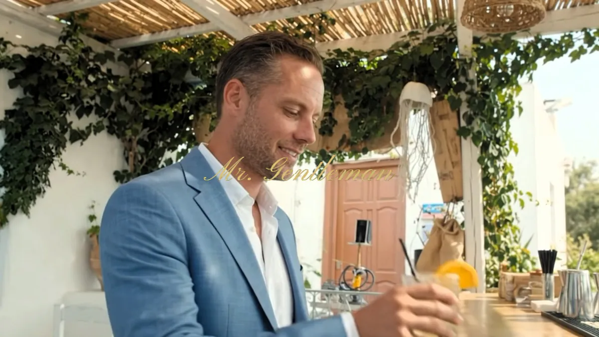 Mr. Gentleman in a light blue blazer with a cocktail at a vine-covered bar in Plaka, Crete
